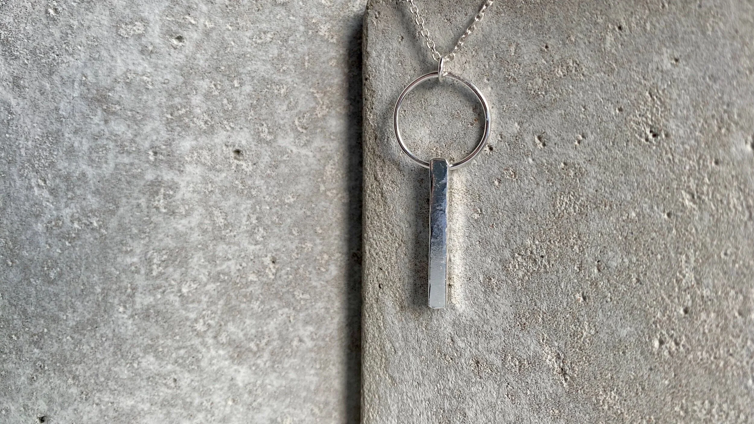 A silver necklace with a circular pendant and a vertical bar charm, hanging against a textured concrete background.
