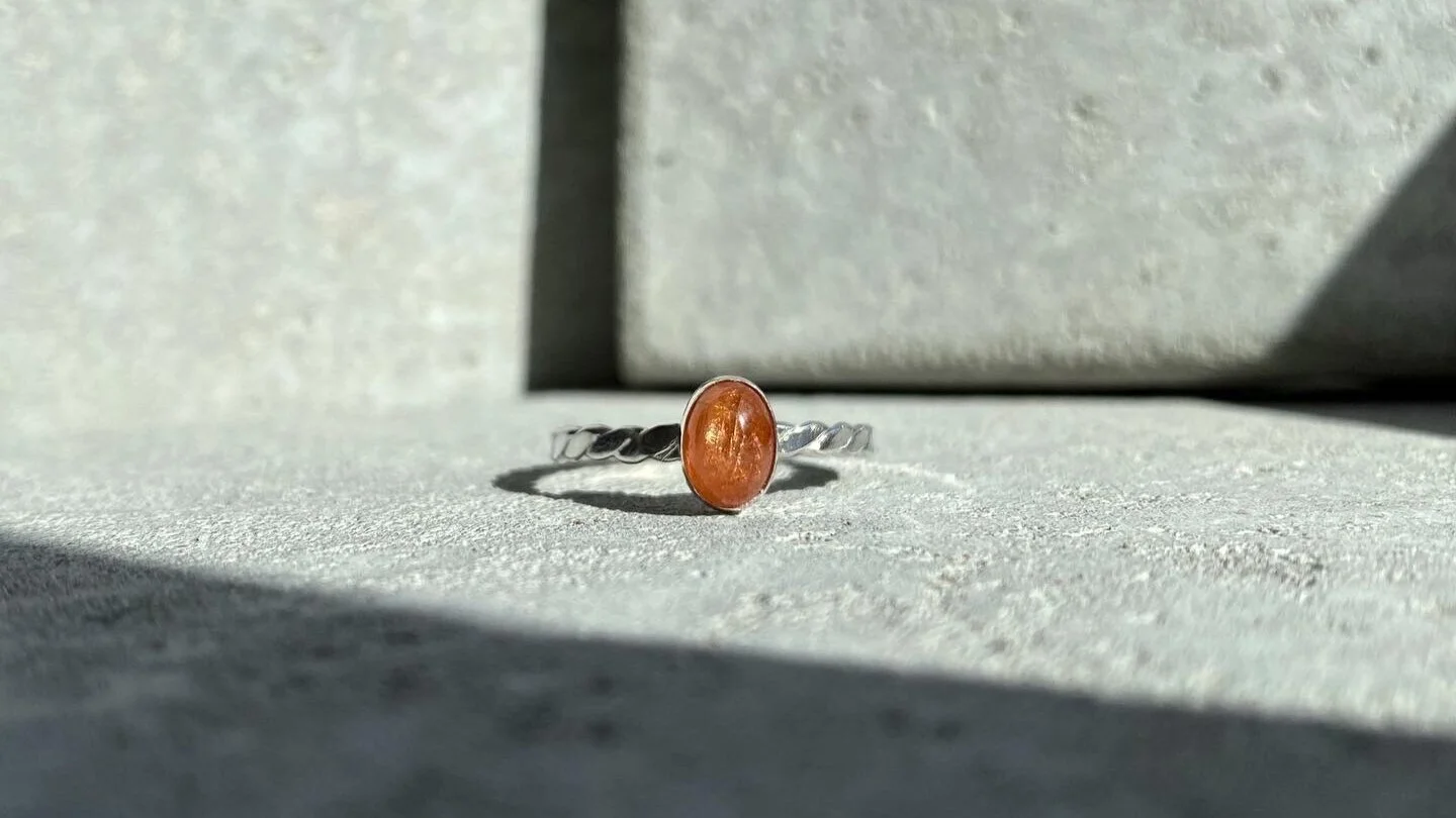 A silver ring with an oval orange gemstone, laid on a textured gray surface with shadows and a background of plain concrete surfaces.