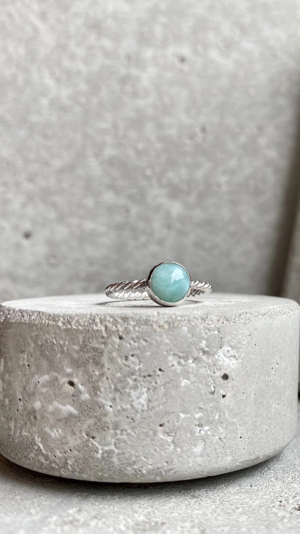 A silver ring with a twisted band and a round turquoise stone, displayed on a textured concrete surface against a gray background.