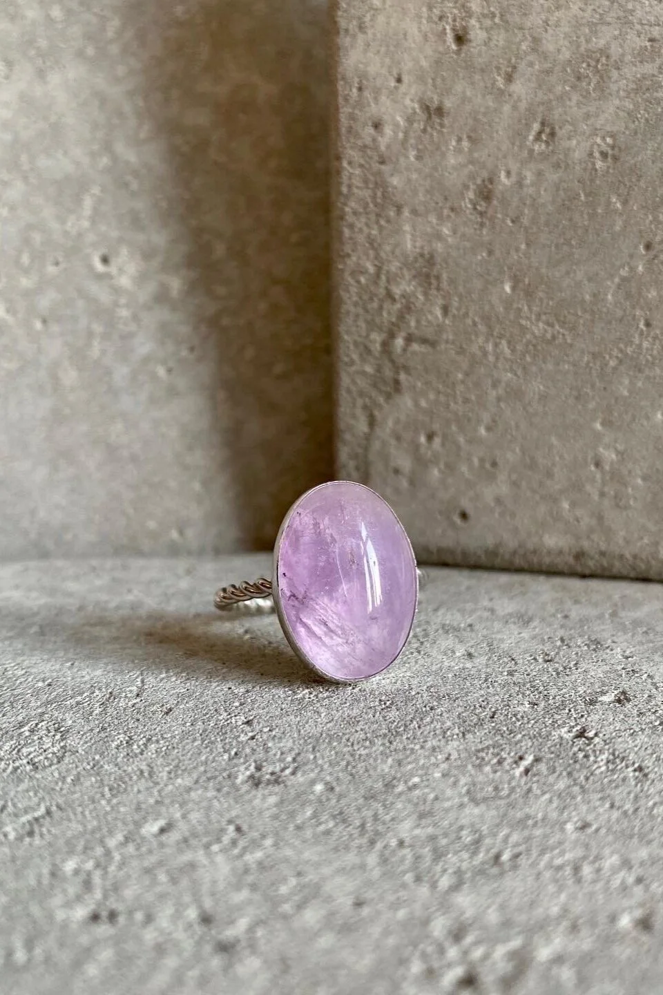 A silver ring with an oval-shaped pinkish-purple gemstone, placed on a textured concrete surface with a concrete wall behind it.