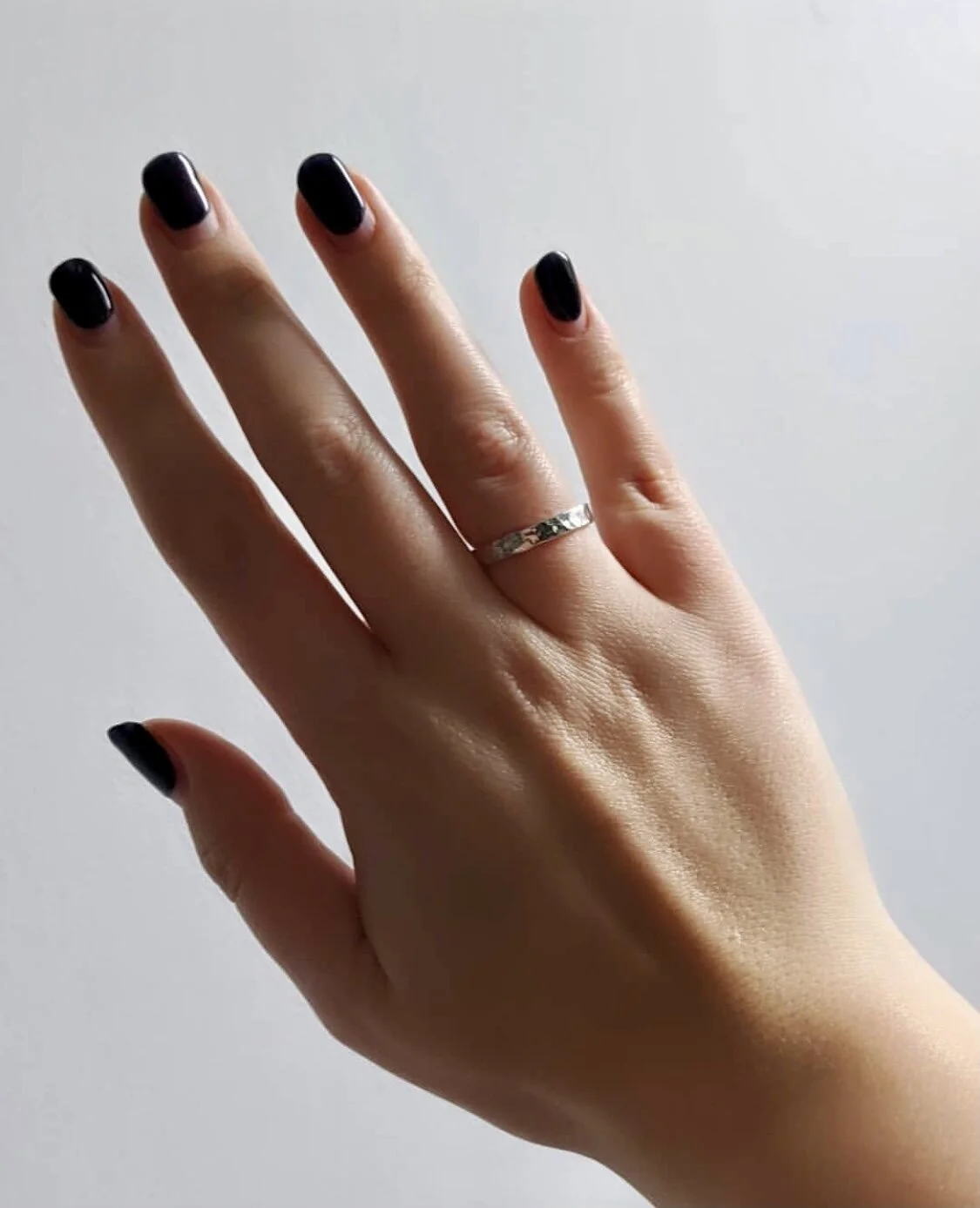 A person's hand with dark nail polish, wearing a silver ring on the middle finger, against a plain light background.
