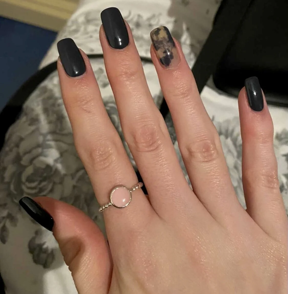 Hand with black manicured nails and a pink ring on the ring finger, against a floral-patterned background.
