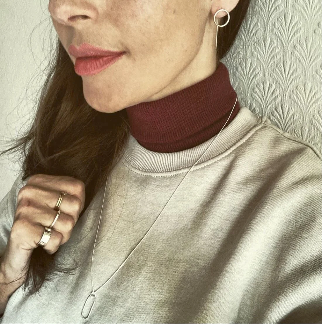Close-up of a woman wearing a beige sweatshirt, red turtleneck, silver necklace, and hoop earrings, with rings on her fingers, and holding a lock of her hair.