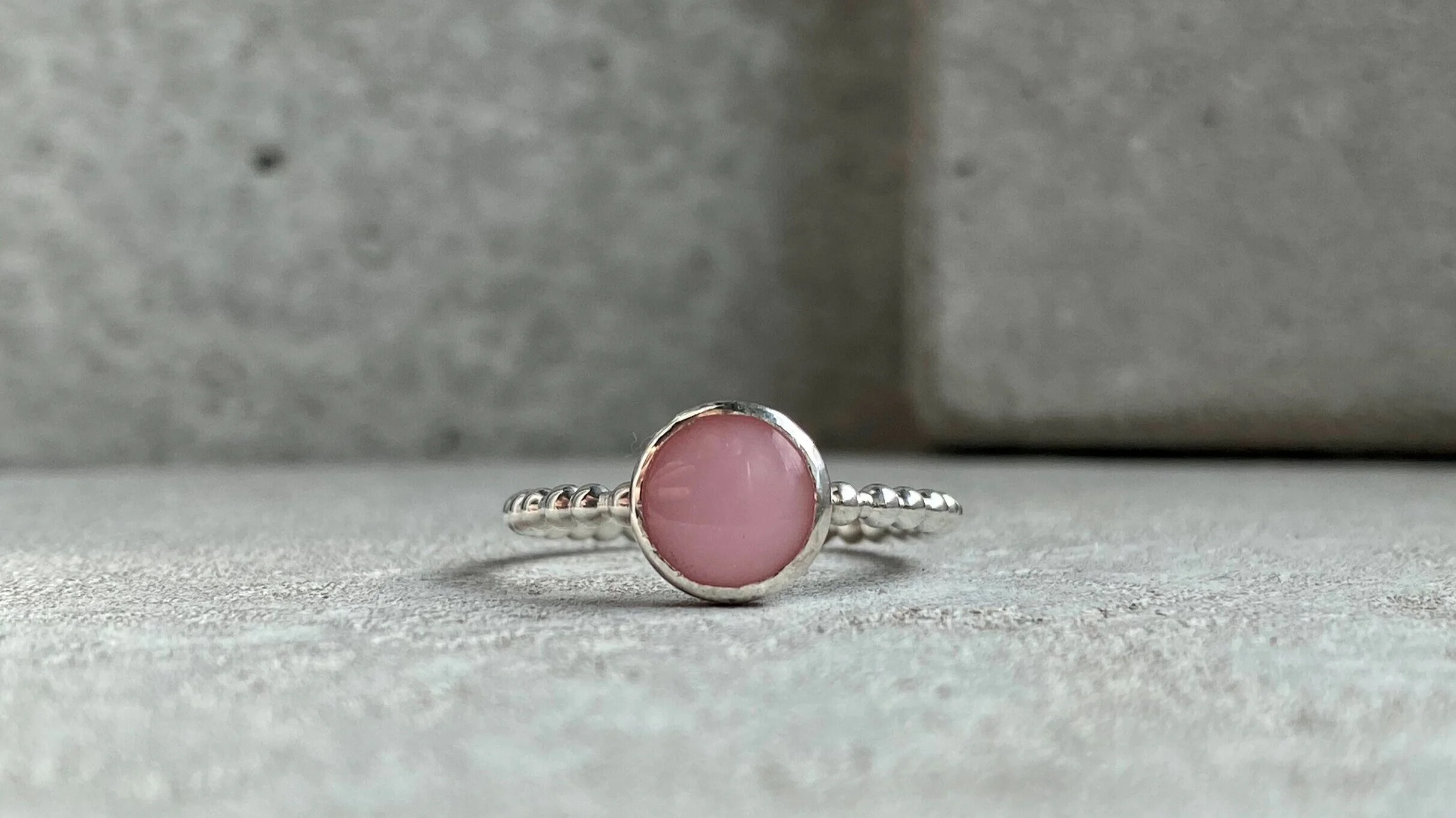 A silver ring with a pink round gemstone in a bezel setting, placed on a gray textured surface with a gray background.