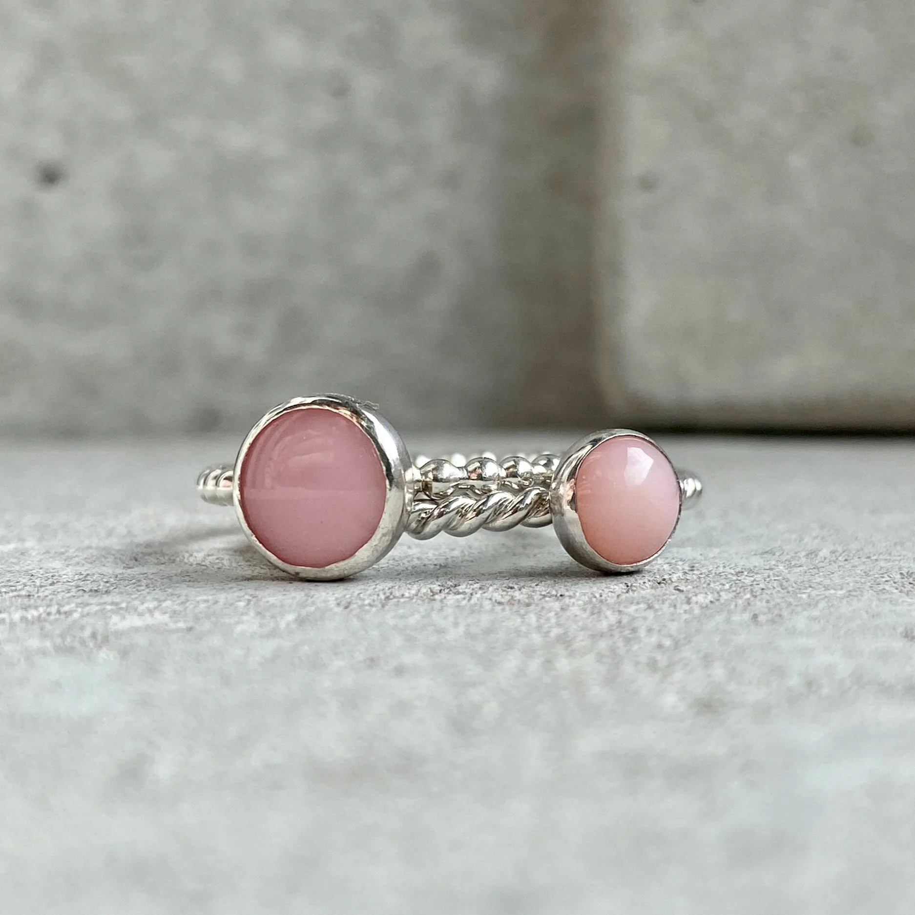 A silver ring with two pink stone accents, one larger and one smaller, on a textured gray surface.