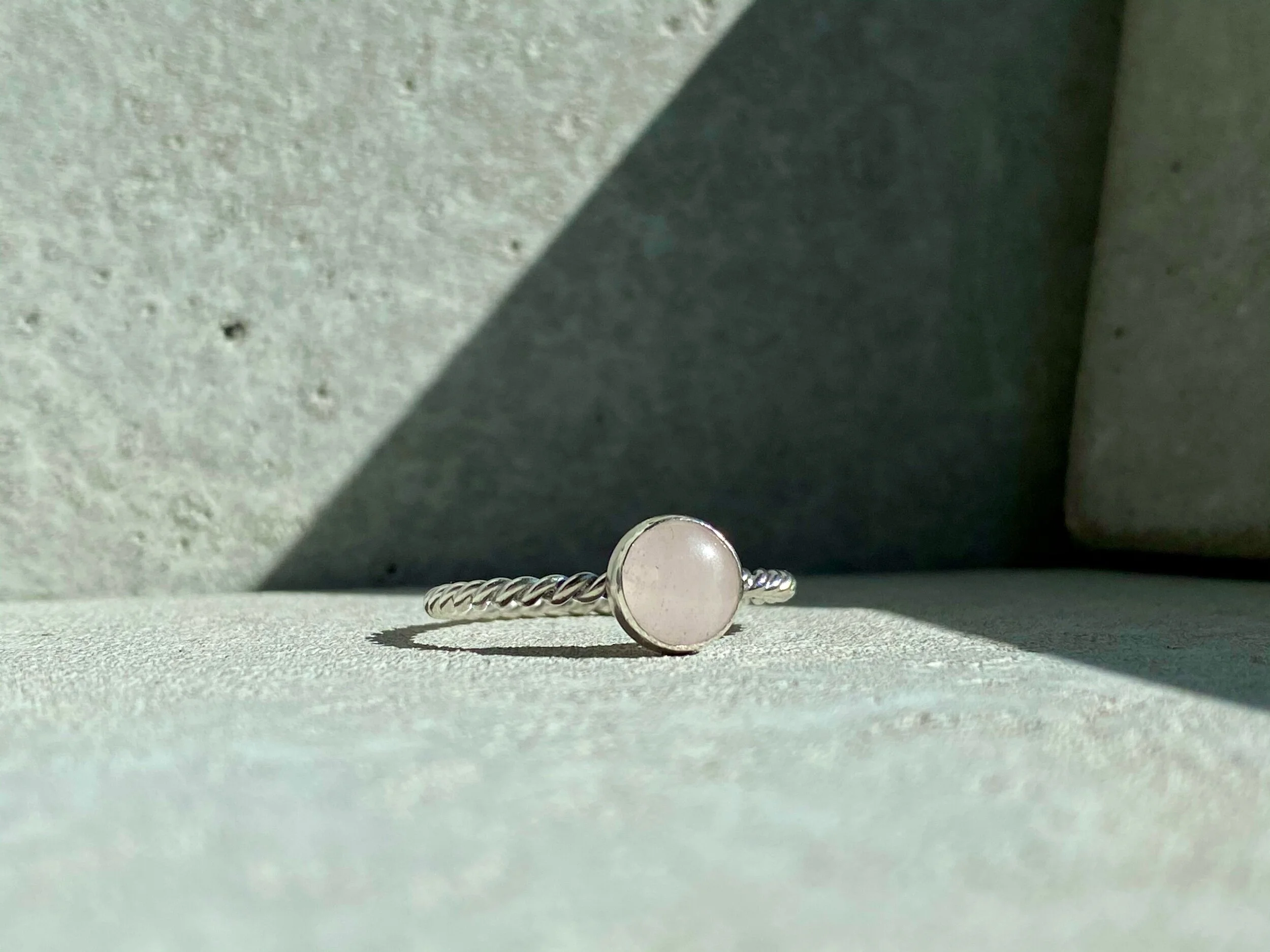 A silver ring with a round, opaque gemstone on a textured surface, with shadows cast by sunlight.