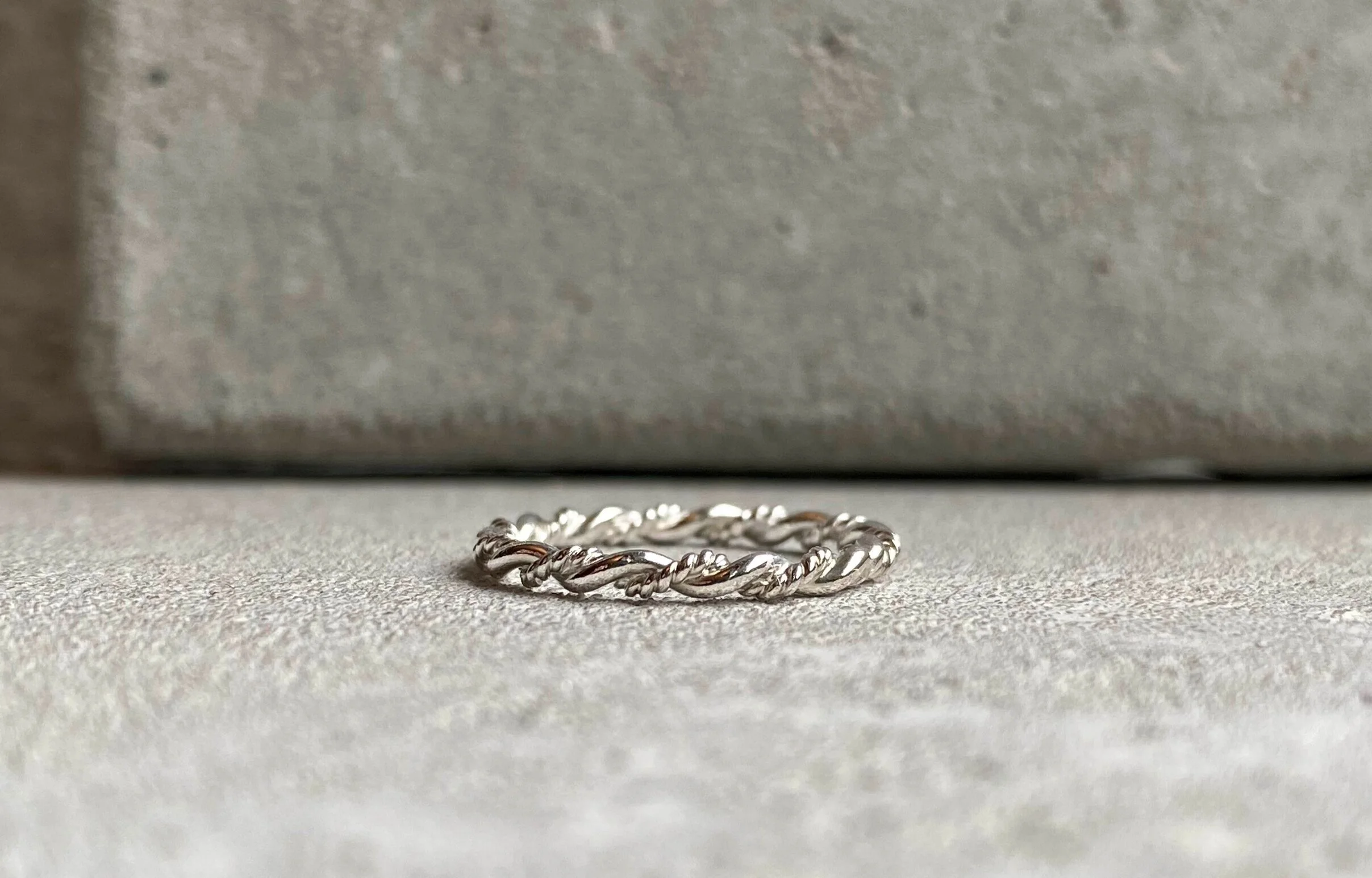 A silver twisted rope-style ring resting on a textured gray surface with a gray box or background in the distance.