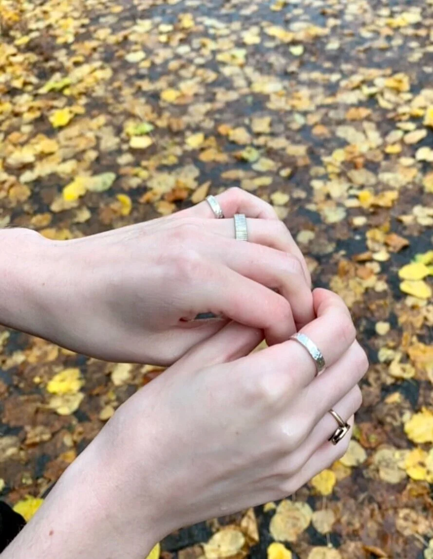Two hands with rings, one with light skin and one with darker skin, holding each other's fingers against a background of fallen yellow leaves.