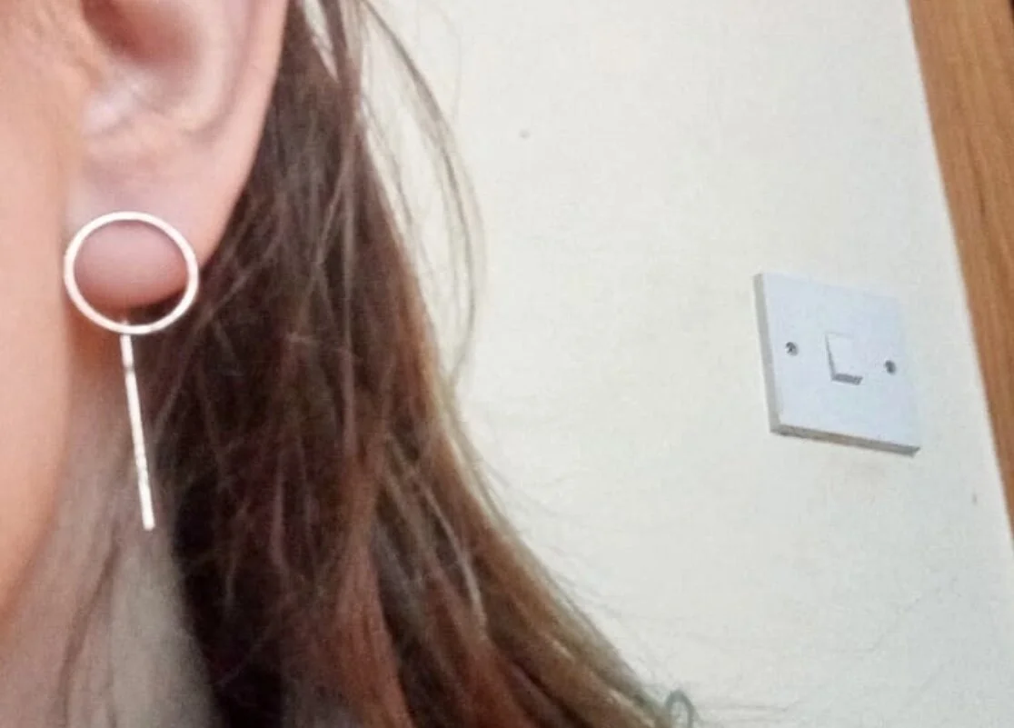 Close-up of a woman's left ear wearing a modern, minimalist silver earring with a circular top and a dangling vertical bar. In the background, a white wall with an electrical switch plate.
