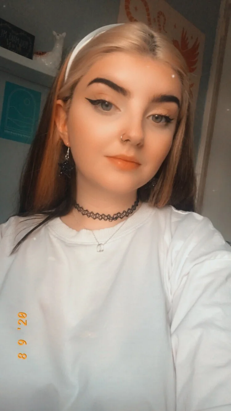 A young woman with blonde and brown hair, wearing a white headband, black choker necklace, earrings, and a white shirt, taking a selfie indoors.