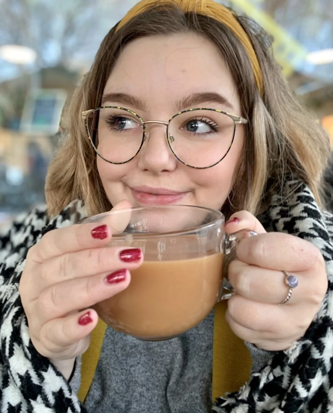 Young woman with glasses and a yellow headband holding a clear glass mug of coffee, smiling softly.