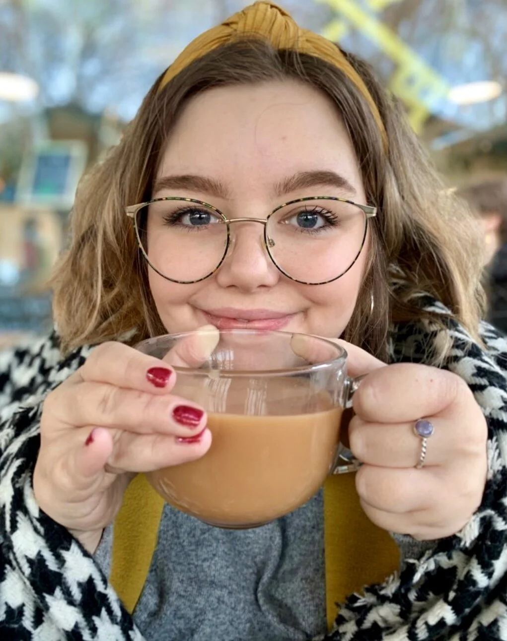 A young woman with glasses and a yellow headband holding a clear glass mug filled with a hot drink, smiling at the camera in a cozy indoor setting.