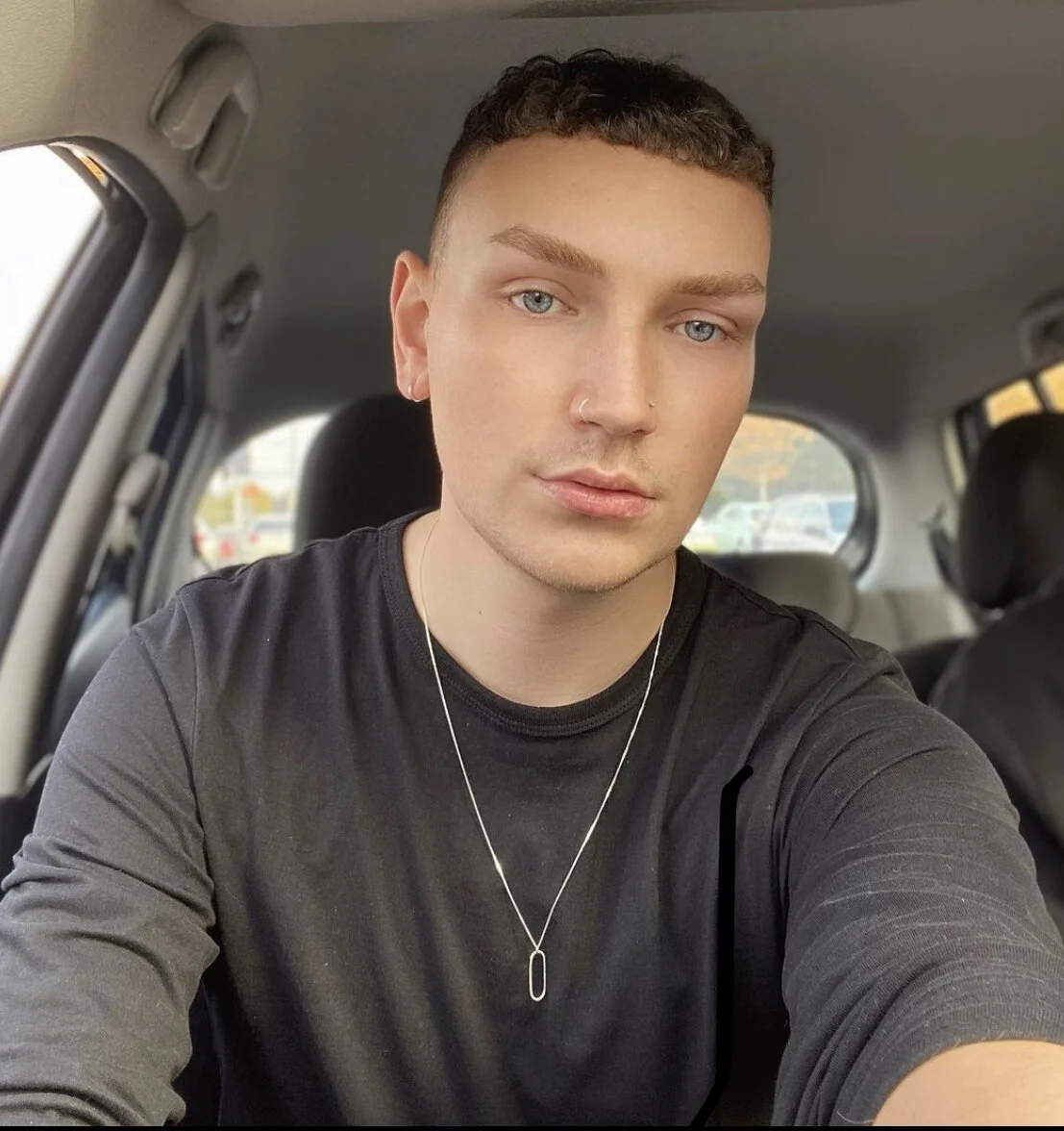 A young man with blue eyes, short dark curly hair, wearing a black shirt, a nose ring, and a necklace, sitting inside a vehicle.