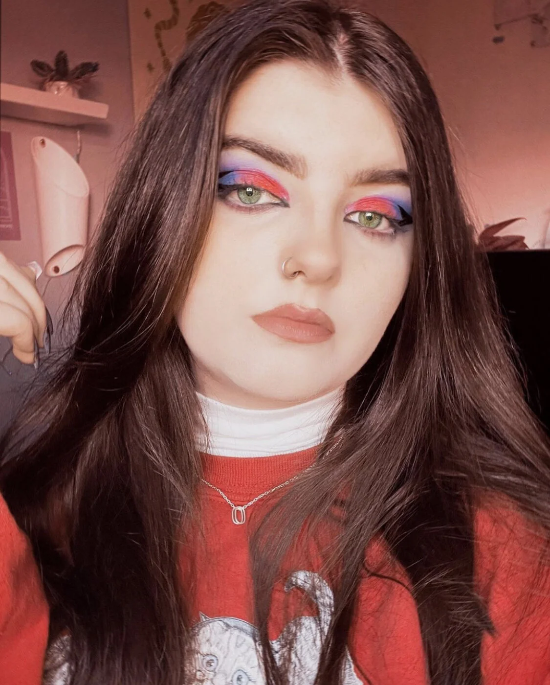 Young woman with long brown hair and colorful eye makeup in pinks, blues, and purples, wearing a nose ring, a red shirt, and a silver necklace, indoors with pink walls and decorative items in the background.