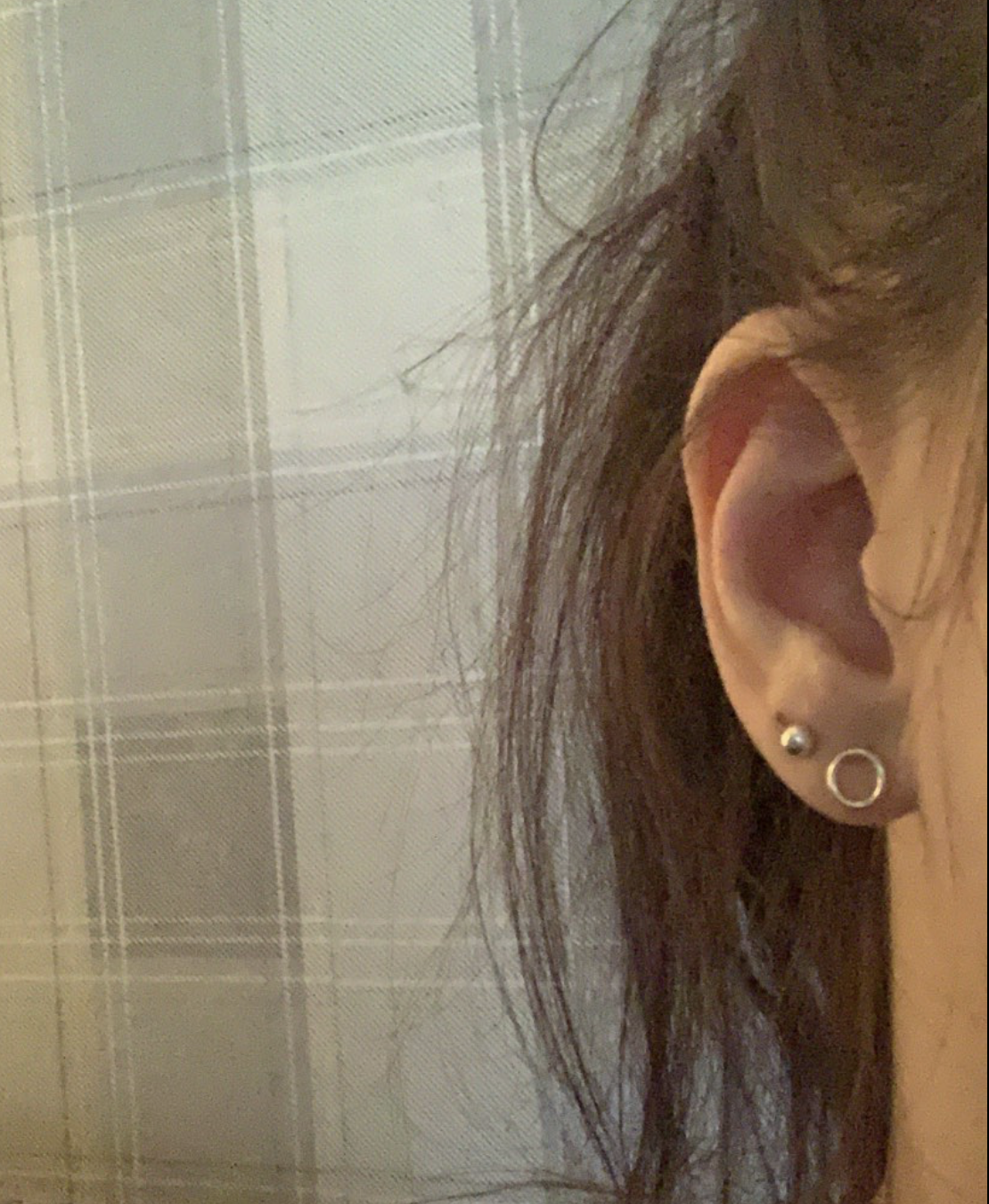Close-up of a person's ear with three earrings: a silver stud, a small silver ball, and a small gold hoop. The background shows a beige plaid pattern.