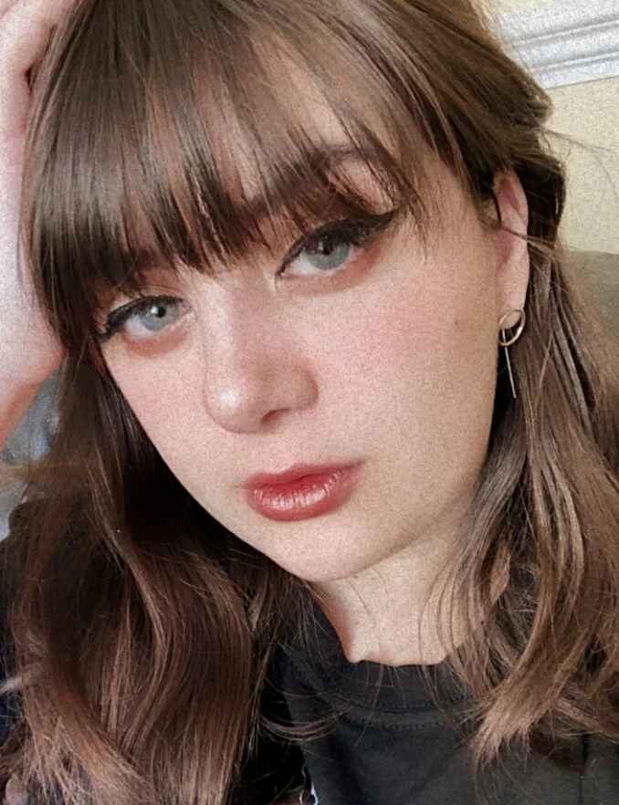 Close-up of a young woman with light brown hair, blue eyes, and red lipstick, wearing hoop earrings, in an indoor setting.