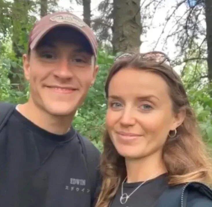 A young man and woman taking a selfie outdoors in a wooded area, both smiling and wearing casual clothing.