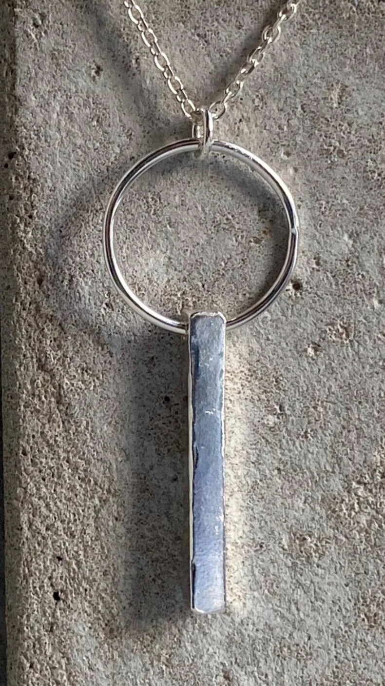 Silver necklace with a pendant in the shape of a vertical rectangular bar, hanging from a chain against a textured concrete background.