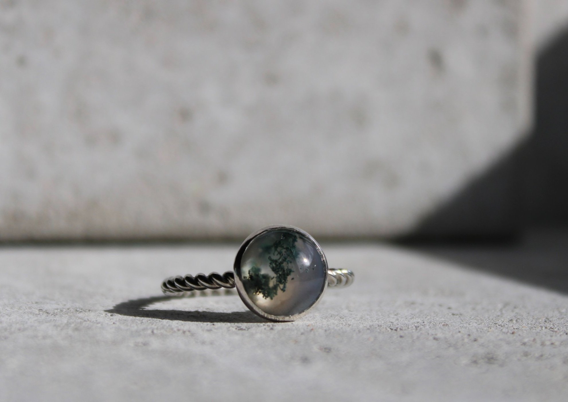 Close-up of a silver ring with a round gemstone featuring a reflective, cloudy interior, resting on a light-colored surface with shadow and background blur.