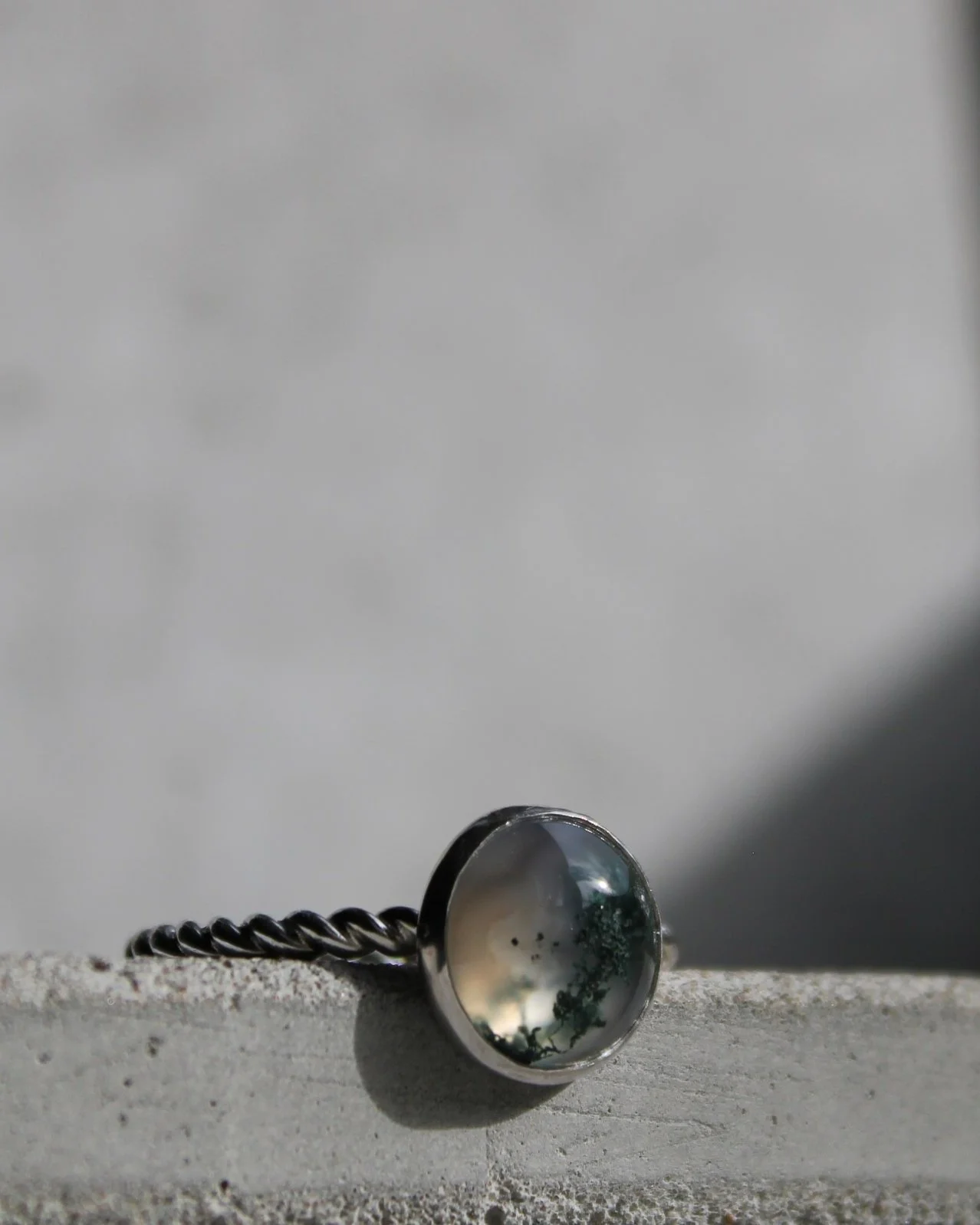 A jewelry ring with a round, dark gemstone set in a silver band, resting on a concrete surface with a blurred background.