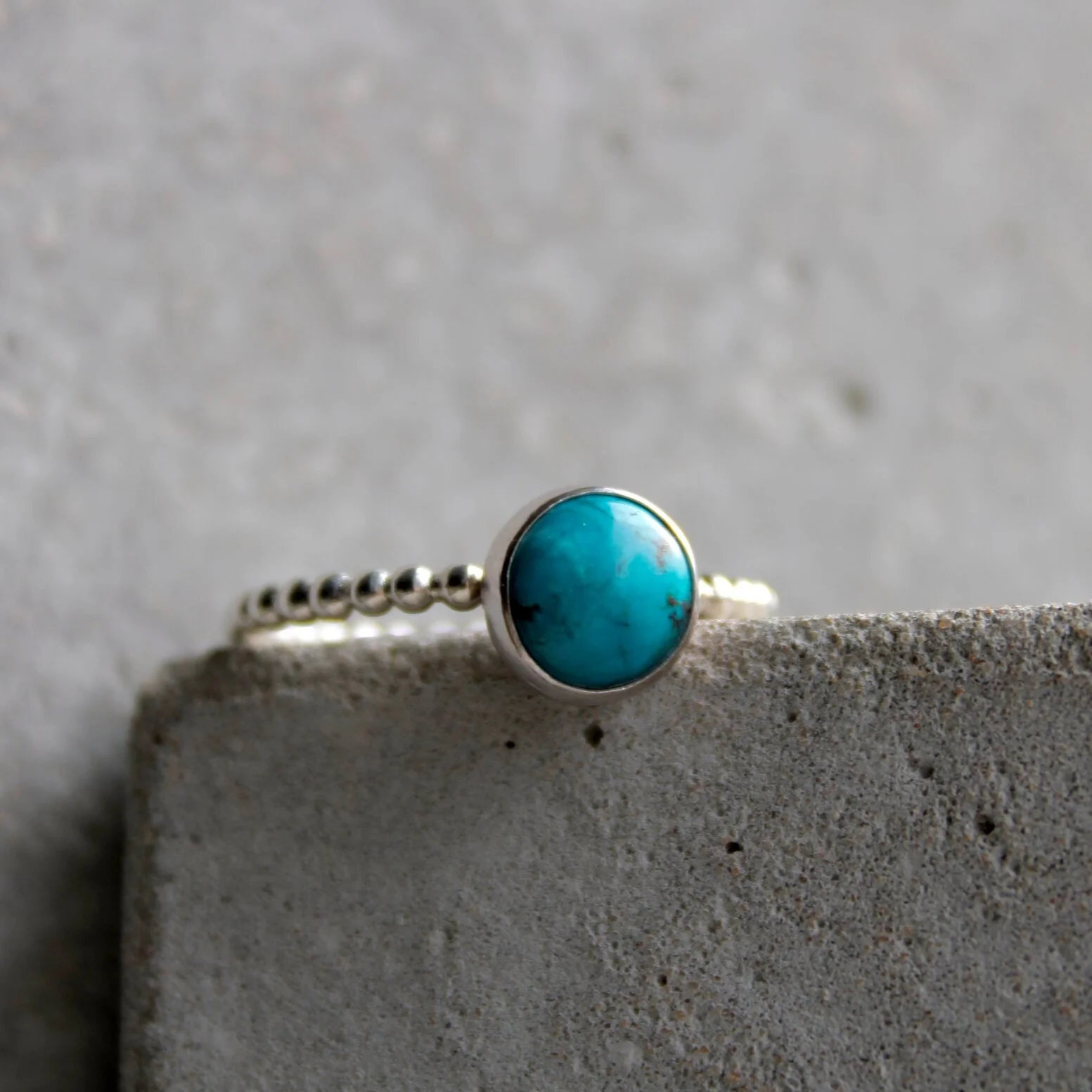 A silver ring with a round turquoise stone, resting on a concrete surface with a plain textured background.