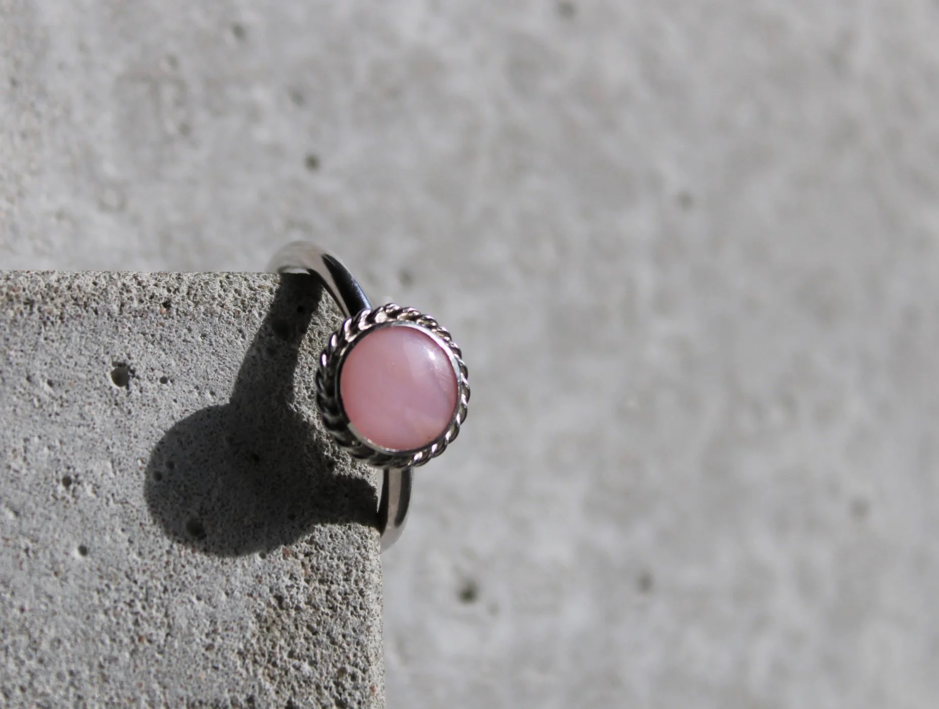 A silver ring with a pink gemstone, resting on a textured gray surface with a shadow cast by the ring.