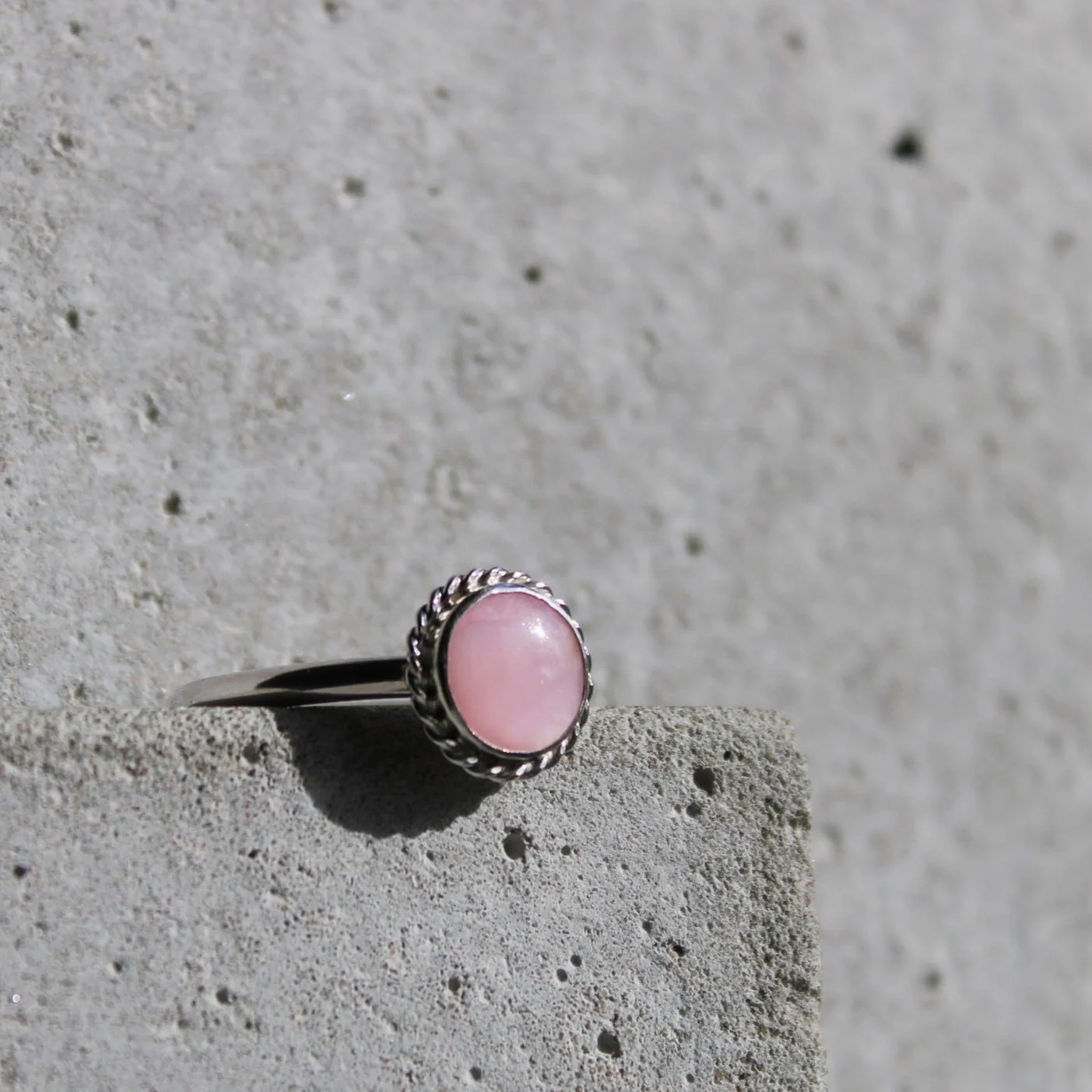 A silver ring with a pink oval gemstone on a rough concrete surface.