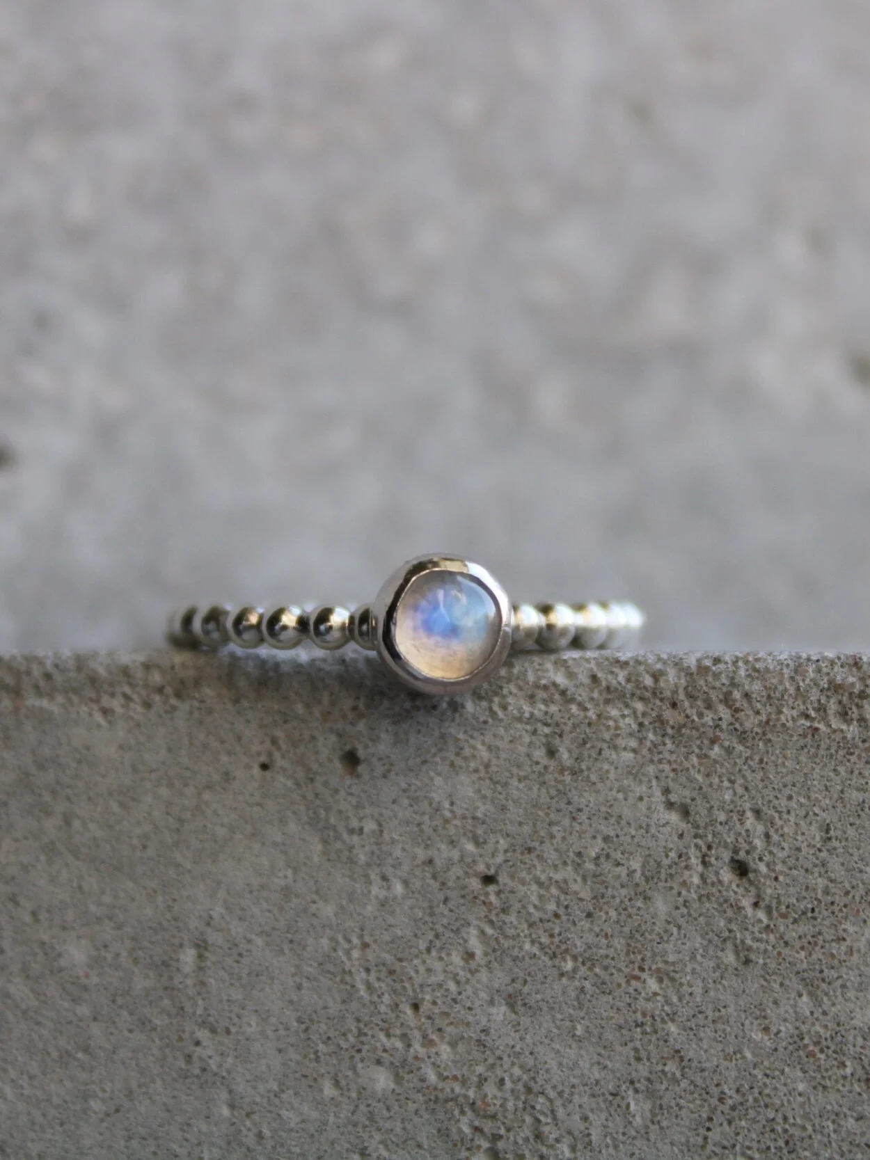 Close-up of a silver ring with a round moonstone in a bezel setting, resting on a concrete surface.