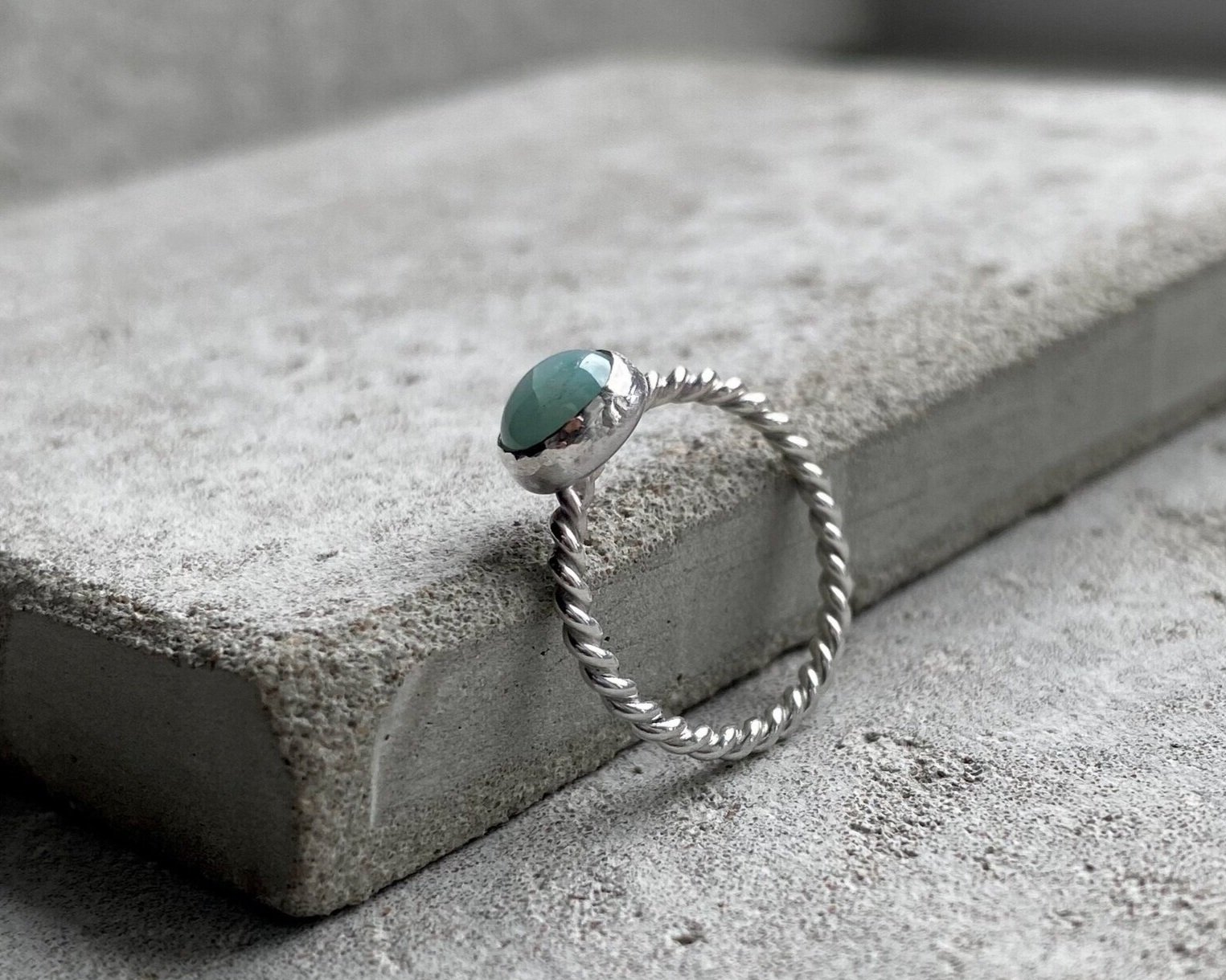 Silver ring with a twisted band and an oval green stone, resting on a concrete surface.