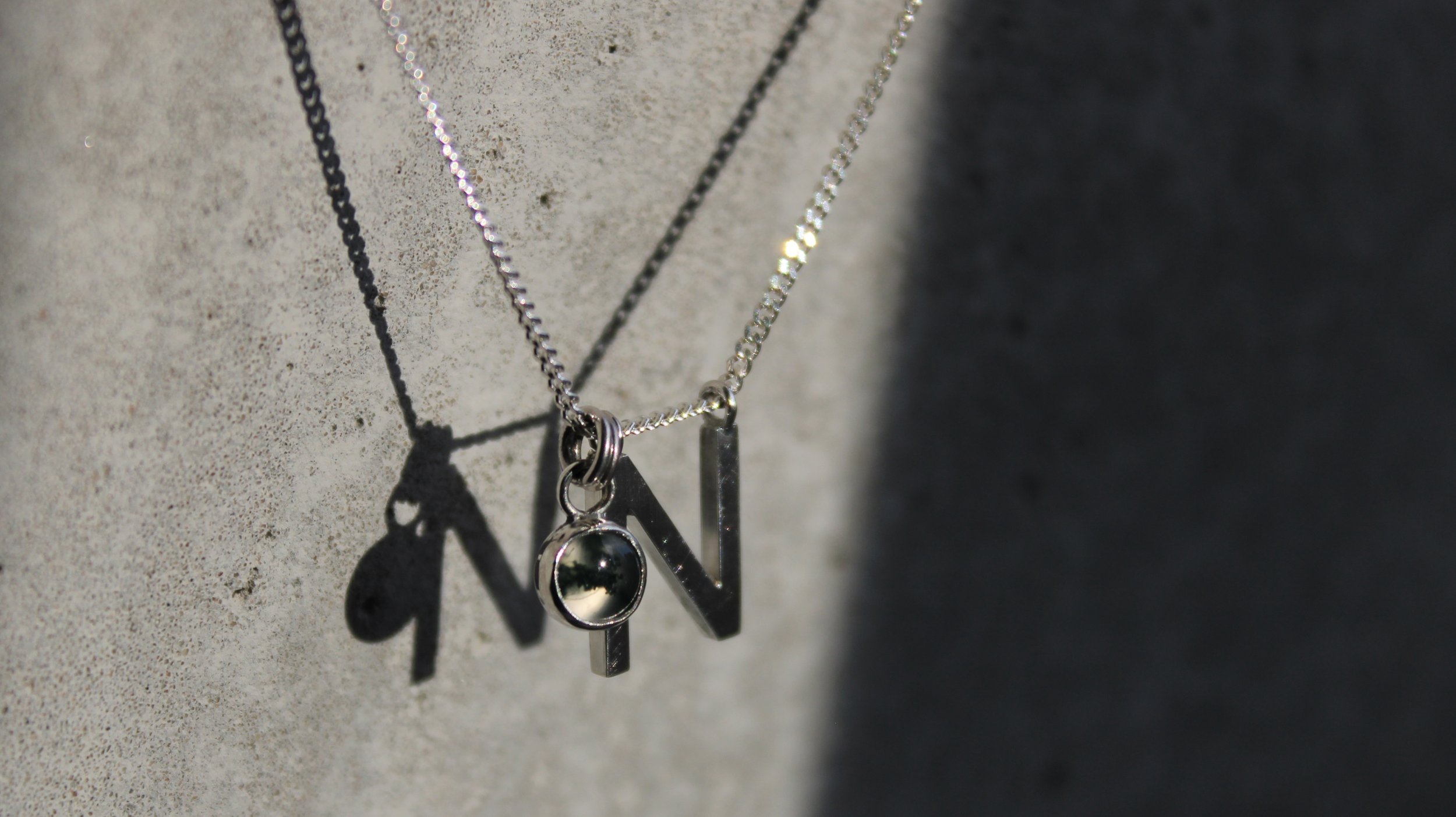Silver necklaces with letter and charm pendants hanging against a textured concrete wall, casting shadows.