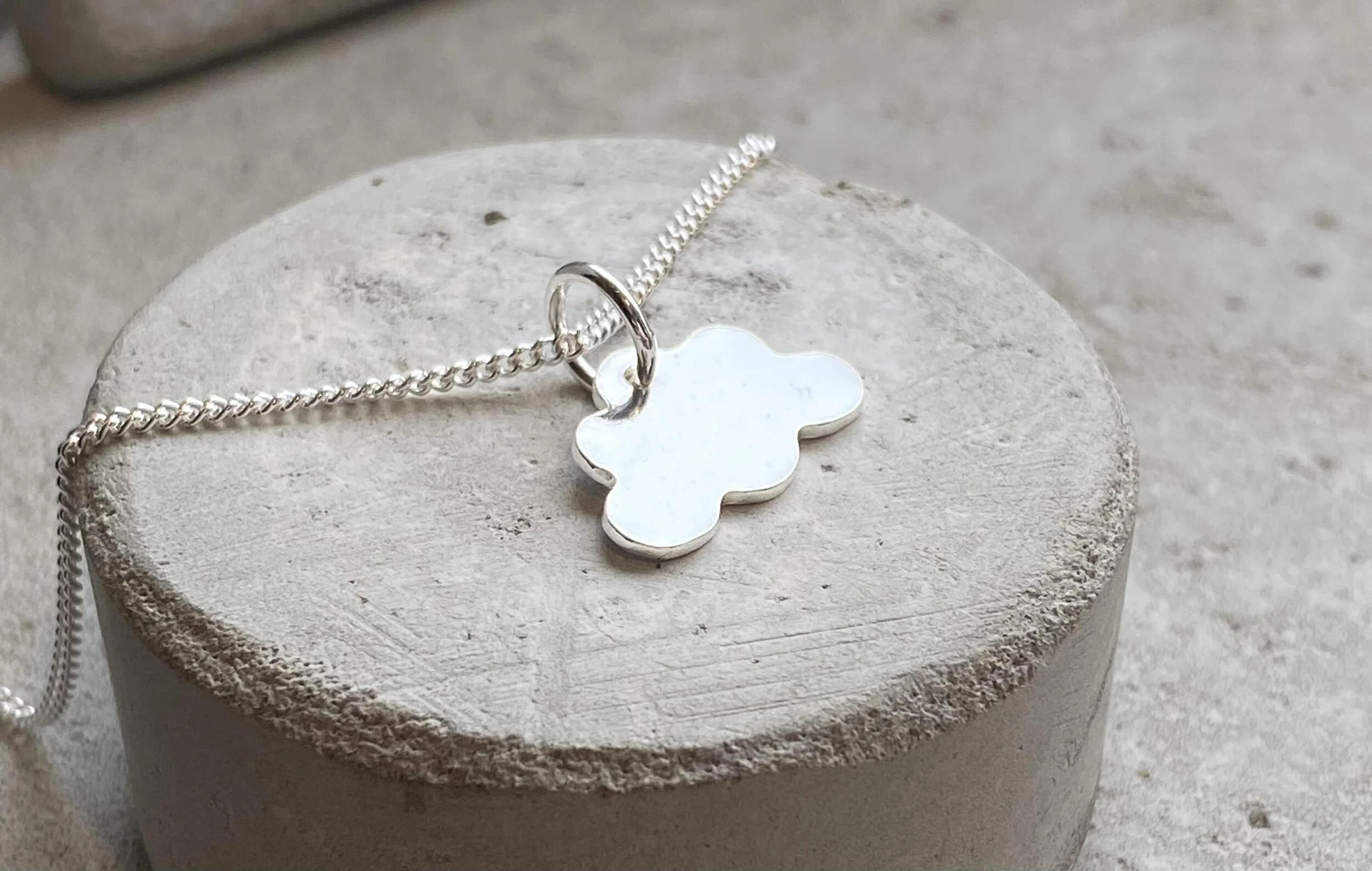 A silver necklace with a cloud-shaped pendant resting on a round, textured, white stone surface.