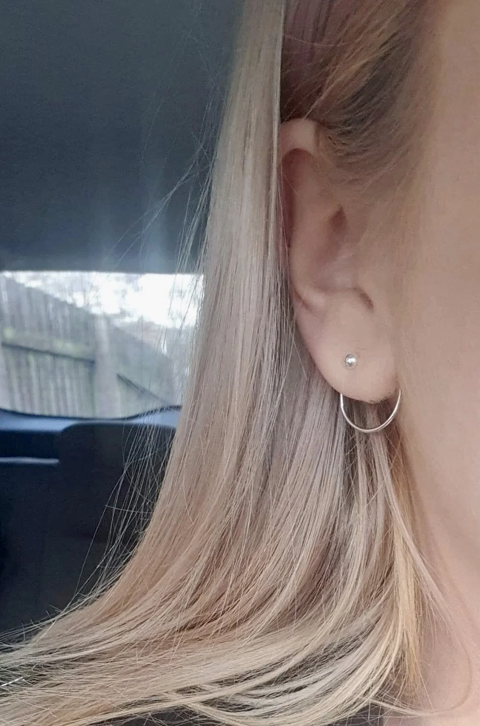 Close-up of a woman's right ear with a small stud earring and a thin hoop earring, and blonde hair partially covering her ear, with a blurred outdoor scene in the background.