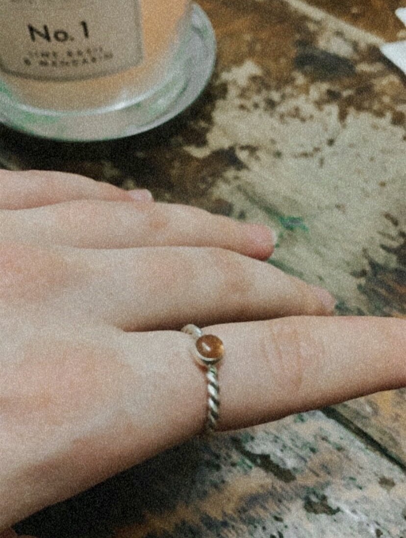 A person's hand wearing a ring with a small amber-colored stone, resting on a wooden surface with a jar in the background.
