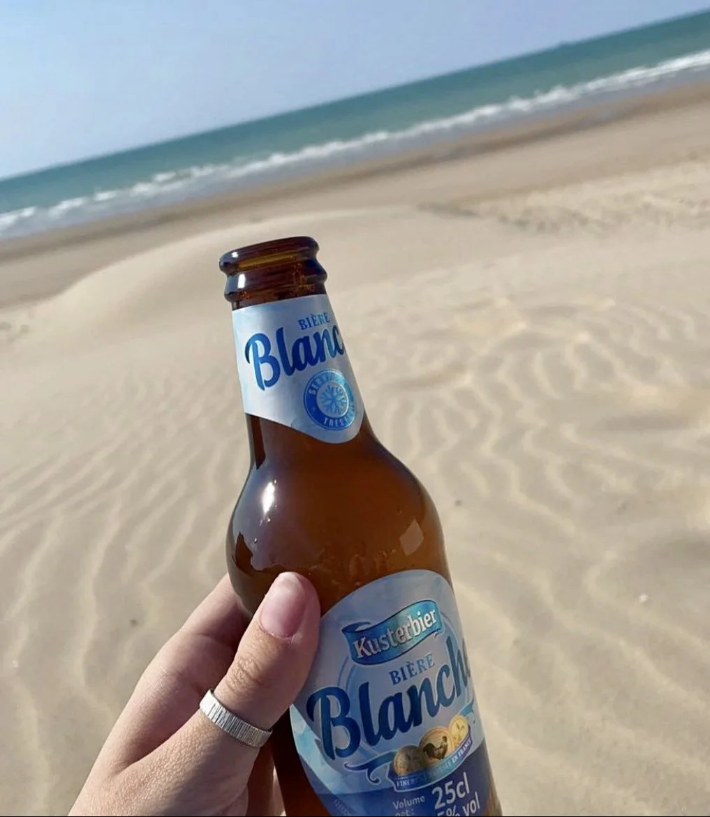 A hand holding a bottle of Kustarbier Biere Blanche on a sandy beach with the ocean and sky in the background.