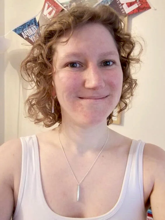 A woman with curly red hair, wearing a white tank top and a silver necklace, smiling indoors with patriotic bunting in the background.