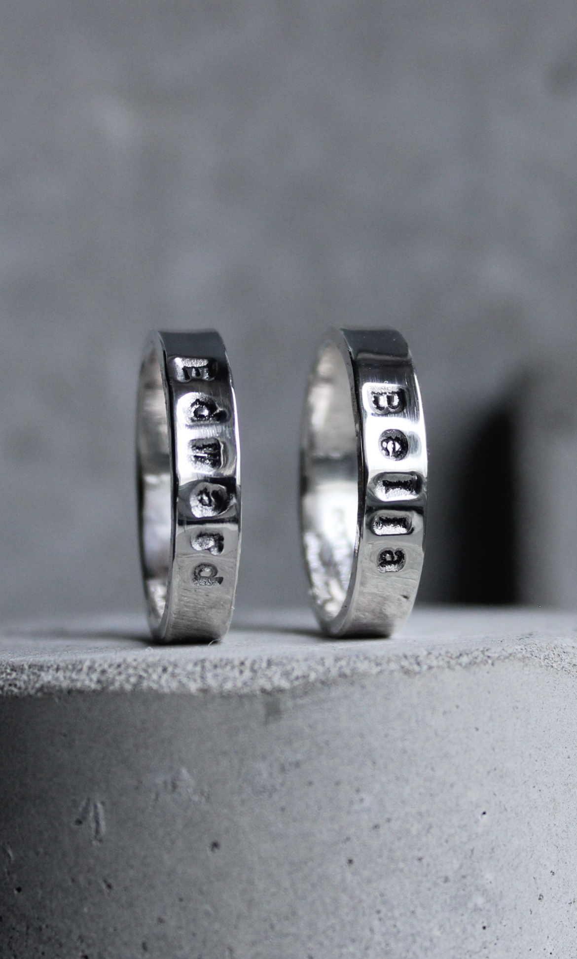 Two silver rings with stamped names, placed on a concrete surface with a gray background.