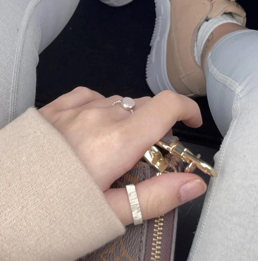 Close-up of a person's hand with rings, holding a set of keys, with legs in jeans and beige sneakers in the background.