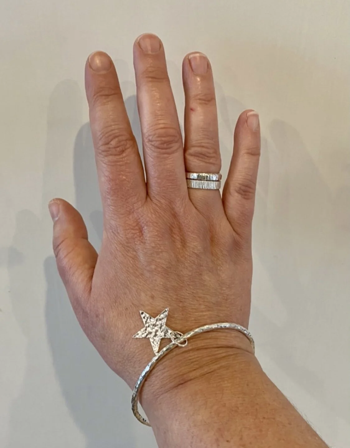 A hand wearing a silver ring, a star-shaped charm bracelet with rhinestones, and a thin silver bangle with rhinestones, against a plain wall background.