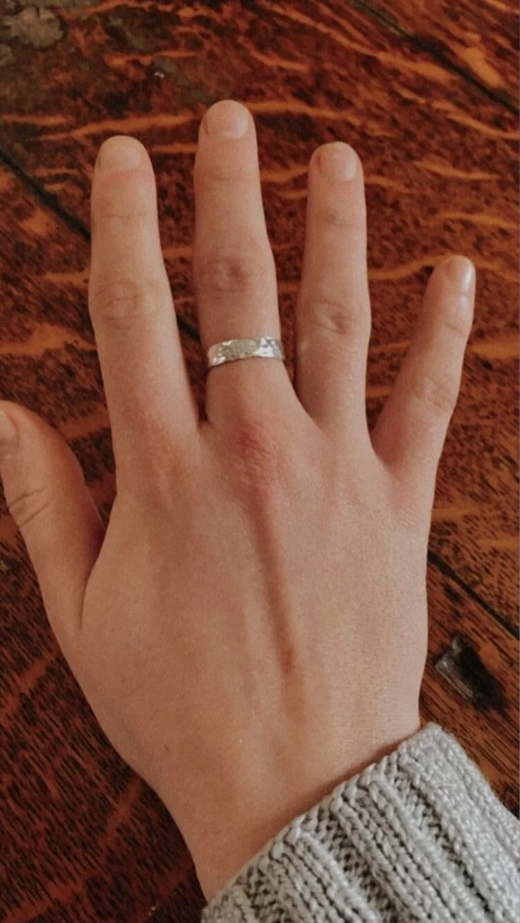 A person's left hand resting on a wooden surface, wearing a silver ring on the ring finger, with a gray knit sweater sleeve visible at the wrist.