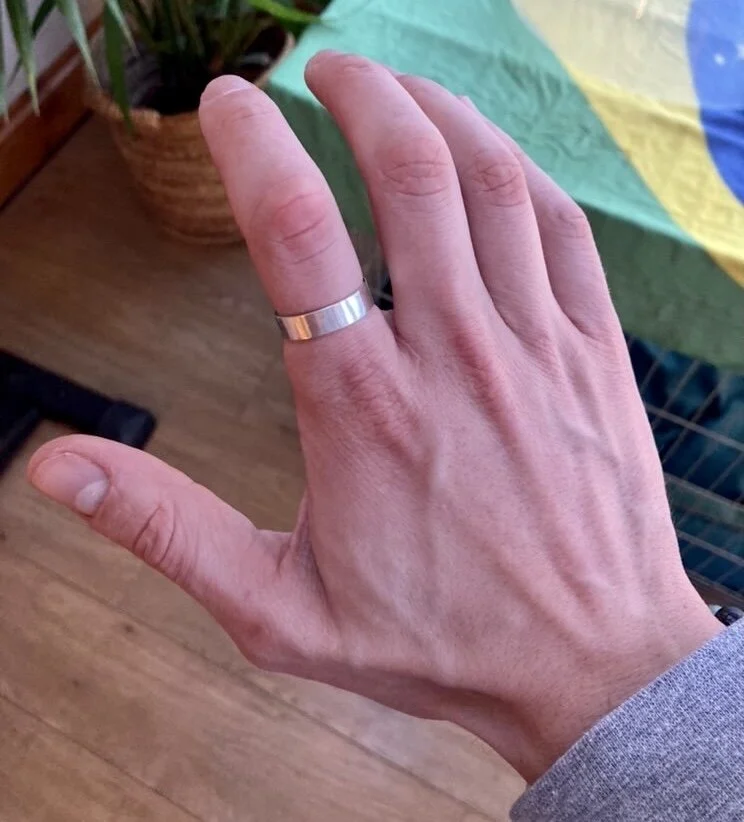Close-up of a person's hand wearing a silver ring on the middle finger, with a background of a green, yellow, and blue item and a wooden floor.