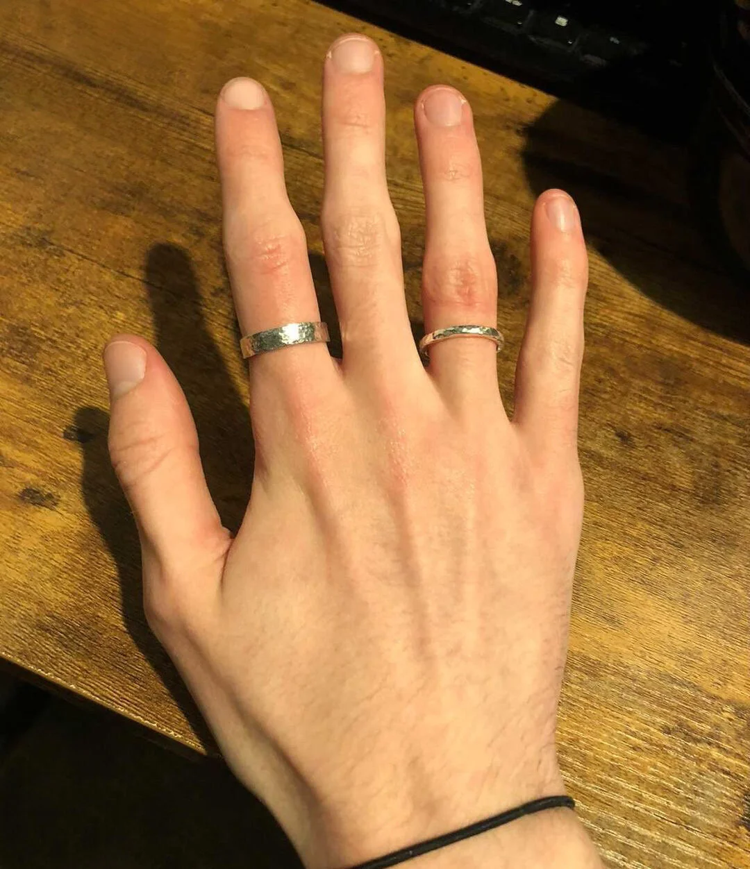 Close-up of a person's left hand resting on a wooden surface, showing two silver rings on the middle and ring fingers, and a black hair tie on the wrist.