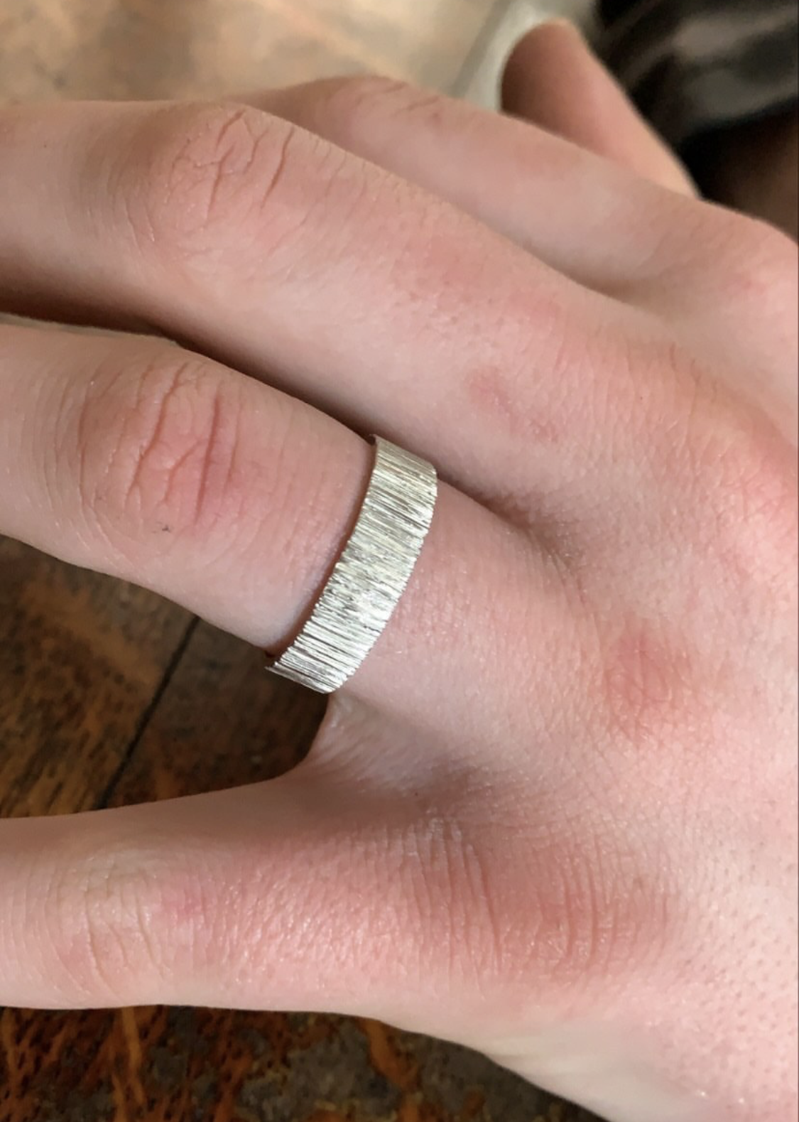 A hand wearing a silver textured ring on the index finger, resting on a wooden surface.