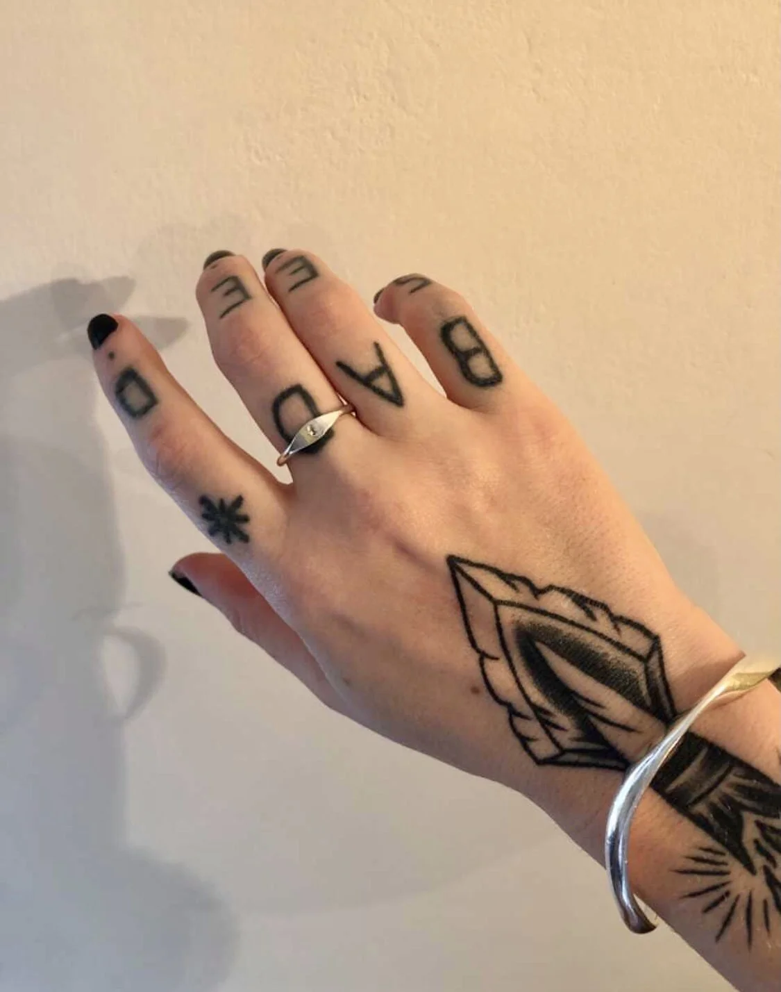 A hand with black nail polish, tattoos, a silver ring on the middle finger, a bracelet, and a tattoo on the wrist. The tattoos include the words "DAD" and "BE," a black star on the index finger, and a geometric design on the wrist.