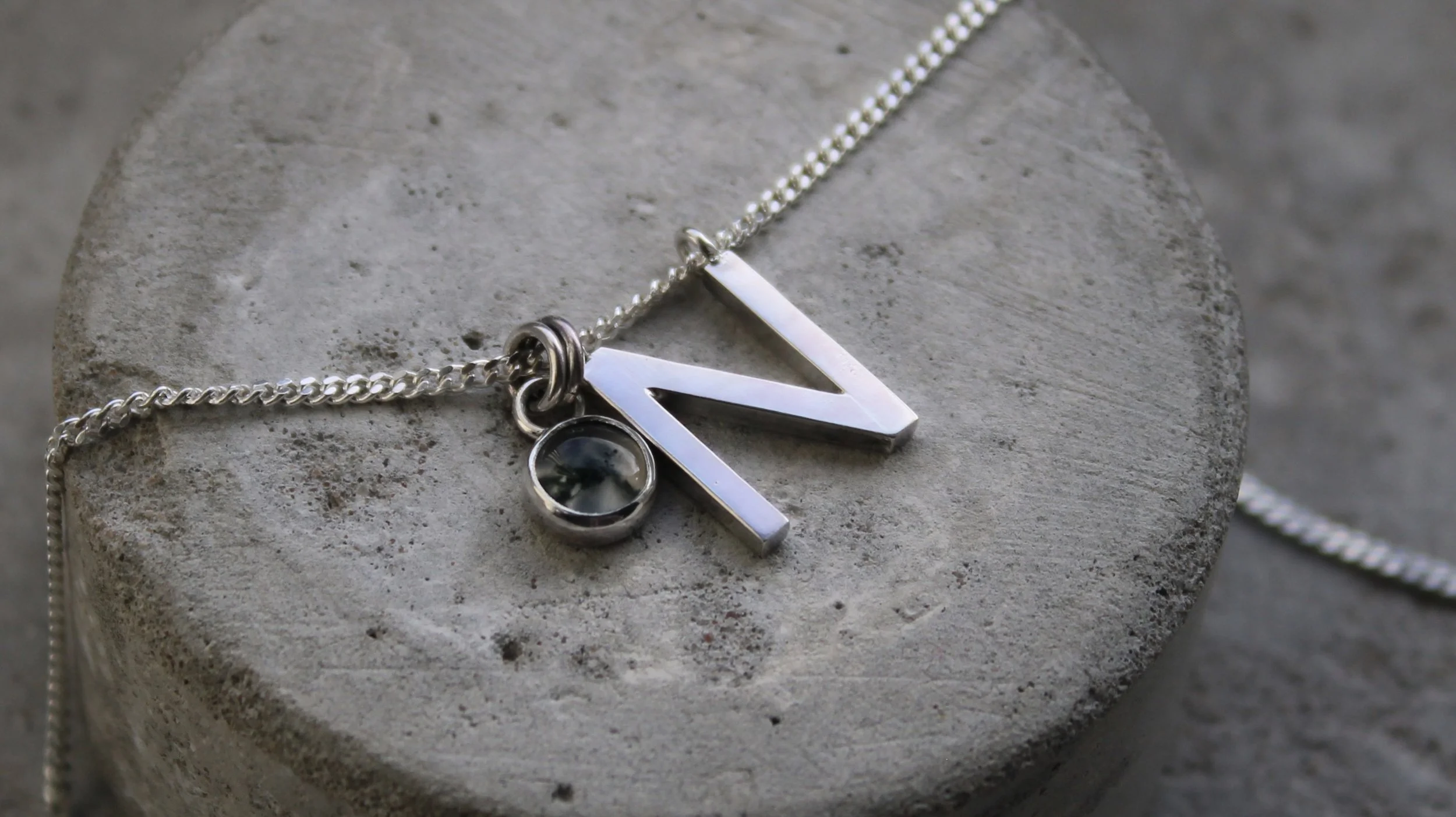 Close-up of a silver necklace with a pendant shaped like the letter 'N' and a small circular gemstone, resting on a rough gray concrete surface.
