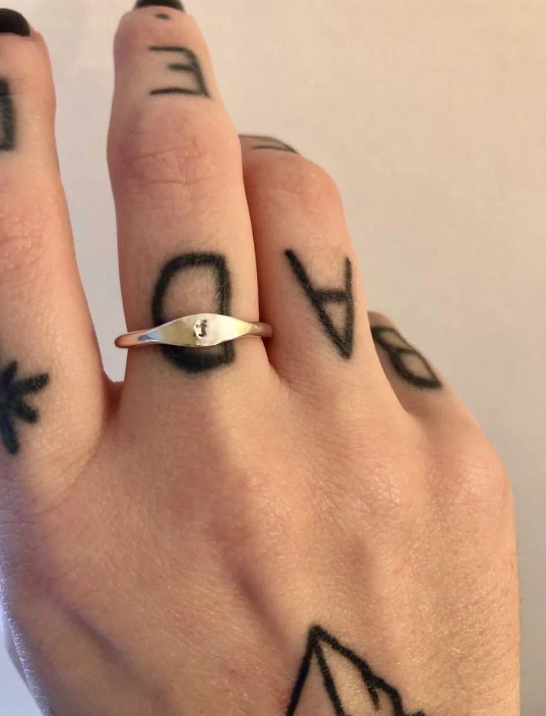 Close-up of a hand with tattoos, wearing a silver ring with an engraved eye symbol, showing black tattooed letters on the fingers.
