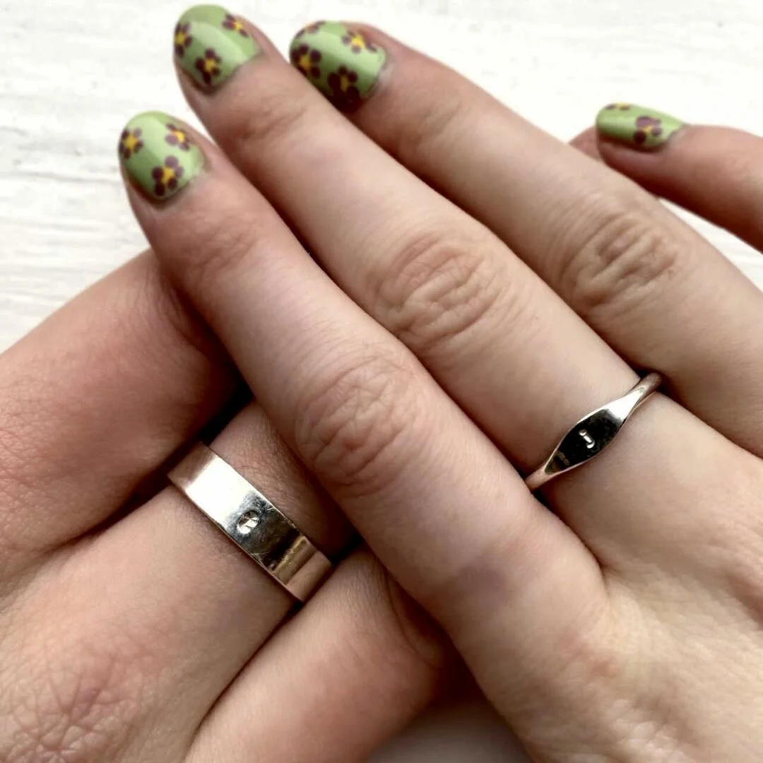 Two hands overlapping, each wearing a silver ring on the ring finger. One hand has colorful floral nail polish with yellow and maroon flowers on a green background.