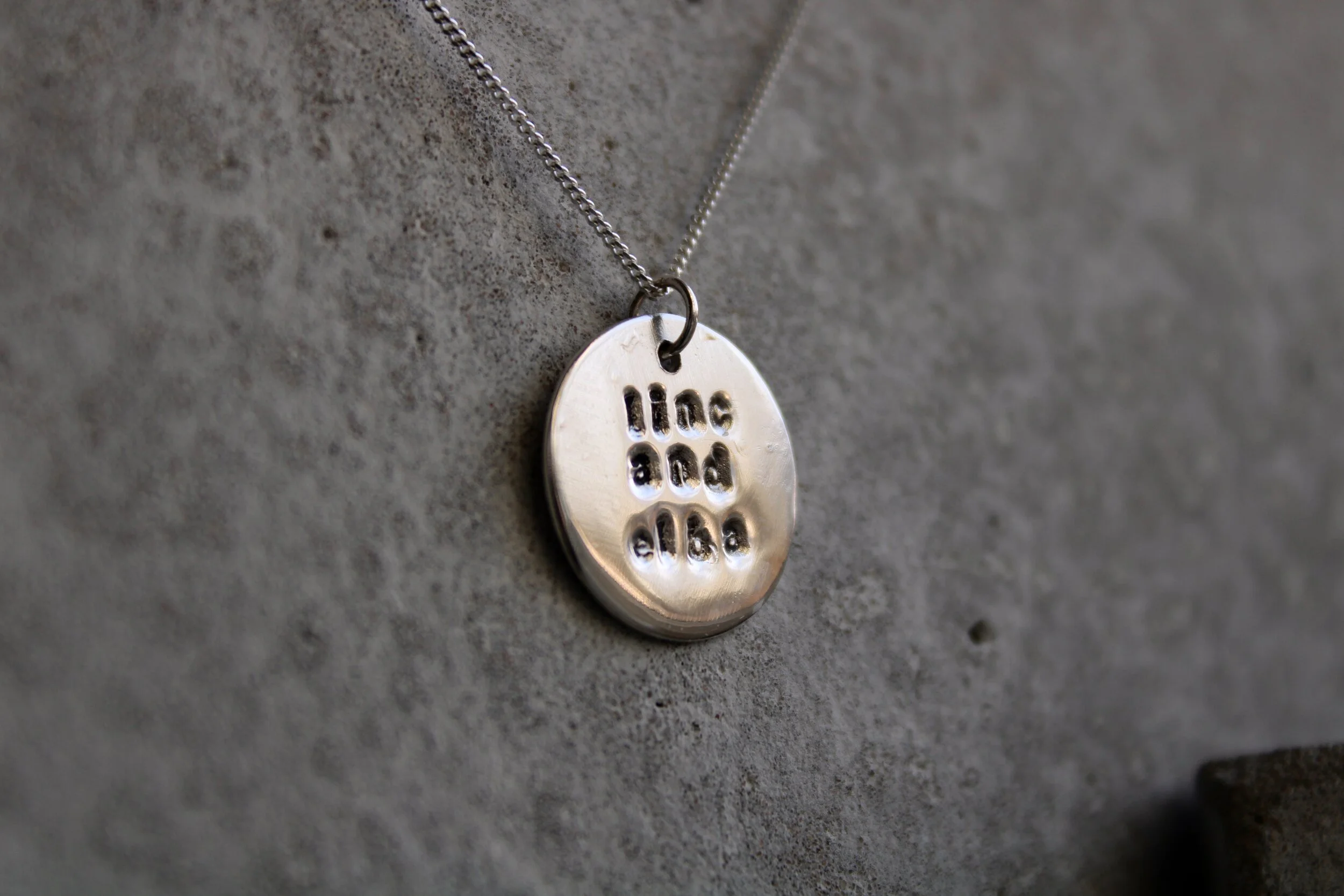 A silver circular necklace pendant. It is hanging on a thin chain against a textured grey background.