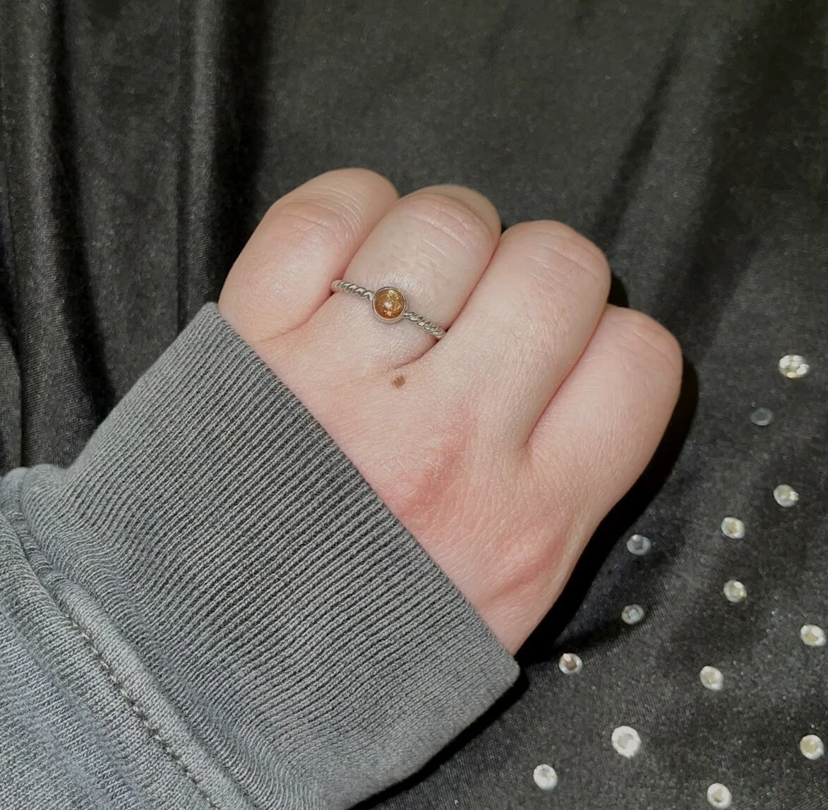 A person's hand with a silver ring featuring a small amber-colored gemstone, resting on a dark fabric background with small shiny embellishments.