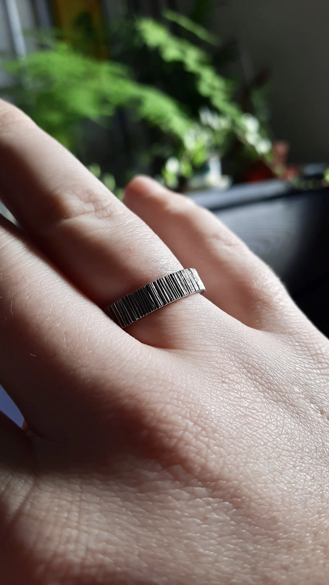 Close-up of a hand wearing a silver ring with vertical ridges, with a background of green plants and blurred objects.