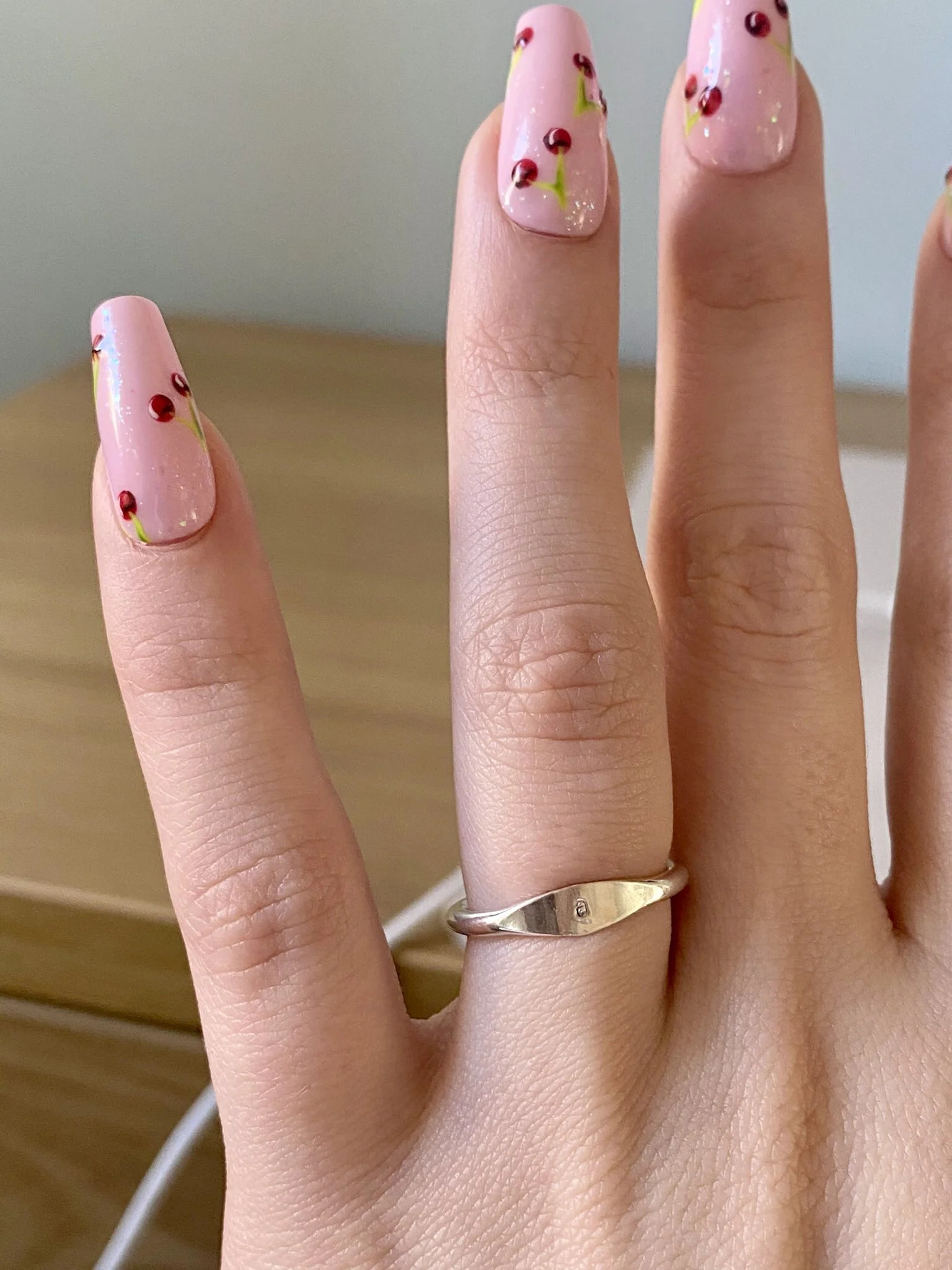 Close-up of a hand showing nails with pink cherry-themed nail art, wearing a silver ring on the ring finger.